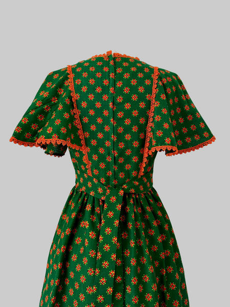 Vintage 1970s boho maxi dress in deep green with orange floral pattern, crochet lace capelet sleeves, waist tie, and colorful border print
