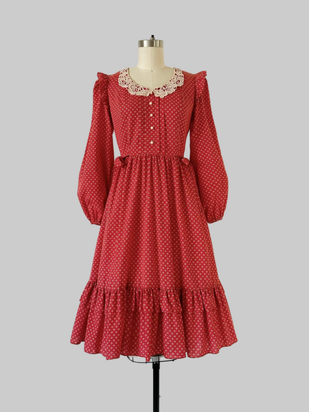 1970s Candi Jones prairie dress in crimson calico print featuring a wide lace collar, ivory rose buttons, self-tie waist bow, ruffled shoulders, and a tiered hem.