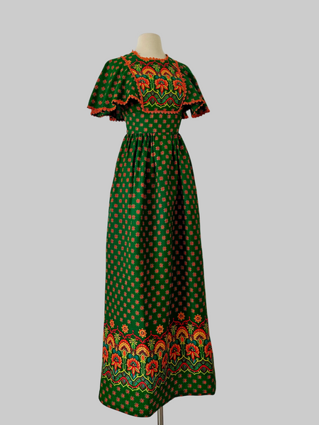 Vintage 1970s boho maxi dress in deep green with orange floral pattern, crochet lace capelet sleeves, waist tie, and colorful border print