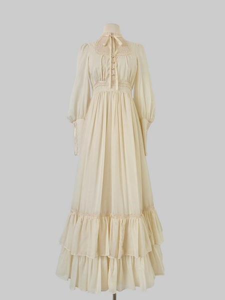 1970s Gunne Sax vintage cream gown with an embroidered yoke, lace-trimmed high neckline with bow tie, faux-pearl buttoned bodice, billowy sleeves with zippered cuffs, waist ties, and a ruffled hem.
