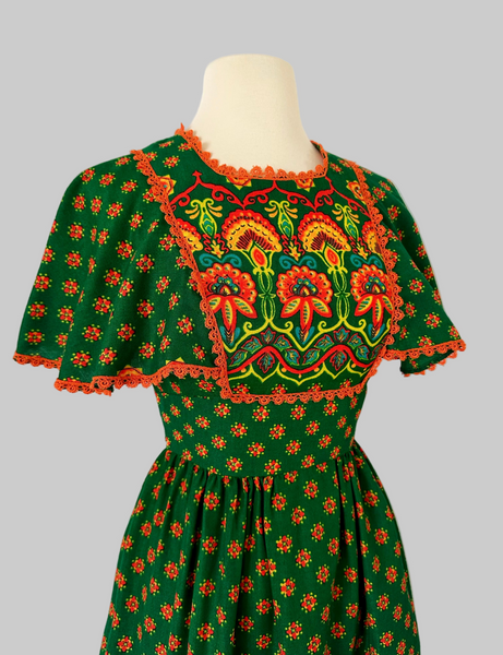 Vintage 1970s boho maxi dress in deep green with orange floral pattern, crochet lace capelet sleeves, waist tie, and colorful border print