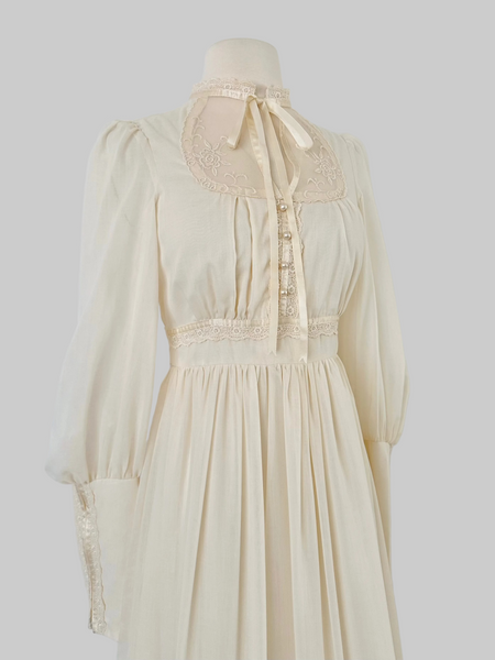 1970s Gunne Sax vintage cream gown with an embroidered yoke, lace-trimmed high neckline with bow tie, faux-pearl buttoned bodice, billowy sleeves with zippered cuffs, waist ties, and a ruffled hem.