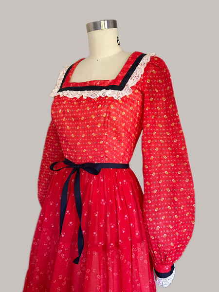 Close Up Mid-1970s crimson red calico prairie maxi dress with off-white lace trim, navy ribbon accents, long sleeves, and a tiered full-length skirt.