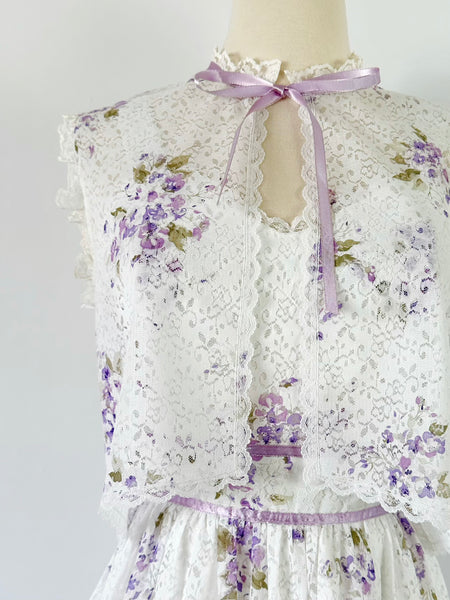 1970s white lace maxi dress with matching cropped jacket, featuring an allover lilac watercolor floral print, sweetheart neckline with scalloped lace trim, wide straps, lavender ribbon details, and a full flowing skirt. Fully lined two-piece vintage set