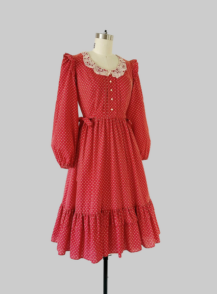 1970s Candi Jones prairie dress in crimson calico print featuring a wide lace collar, ivory rose buttons, self-tie waist bow, ruffled shoulders, and a tiered hem.