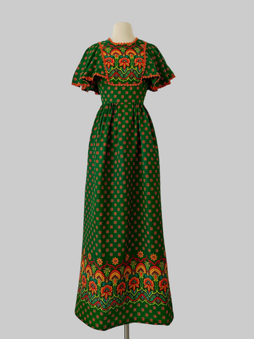 Vintage 1970s boho maxi dress in deep green with orange floral pattern, crochet lace capelet sleeves, waist tie, and colorful border print