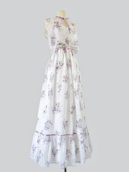 1970s white lace maxi dress with matching cropped jacket, featuring an allover lilac watercolor floral print, sweetheart neckline with scalloped lace trim, wide straps, lavender ribbon details, and a full flowing skirt. Fully lined two-piece vintage set