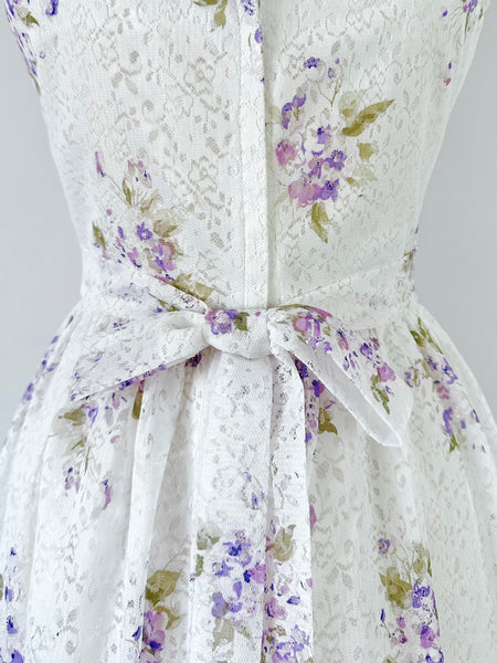 1970s white lace maxi dress with matching cropped jacket, featuring an allover lilac watercolor floral print, sweetheart neckline with scalloped lace trim, wide straps, lavender ribbon details, and a full flowing skirt. Fully lined two-piece vintage set