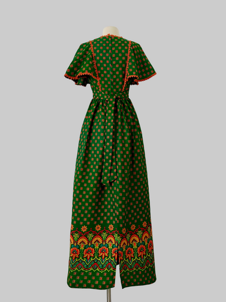 Vintage 1970s boho maxi dress in deep green with orange floral pattern, crochet lace capelet sleeves, waist tie, and colorful border print
