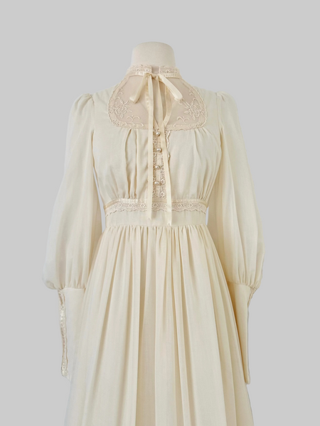 1970s Gunne Sax vintage cream gown with an embroidered yoke, lace-trimmed high neckline with bow tie, faux-pearl buttoned bodice, billowy sleeves with zippered cuffs, waist ties, and a ruffled hem.