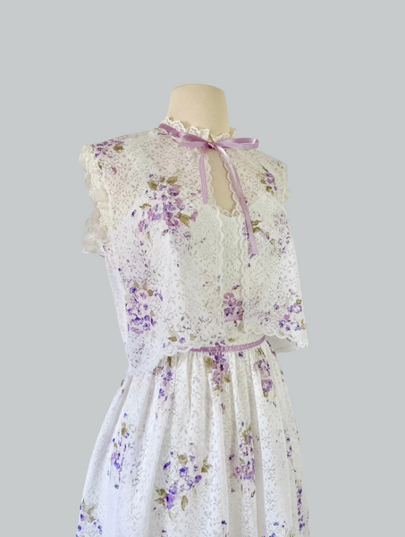 1970s white lace maxi dress with matching cropped jacket, featuring an allover lilac watercolor floral print, sweetheart neckline with scalloped lace trim, wide straps, lavender ribbon details, and a full flowing skirt. Fully lined two-piece vintage set