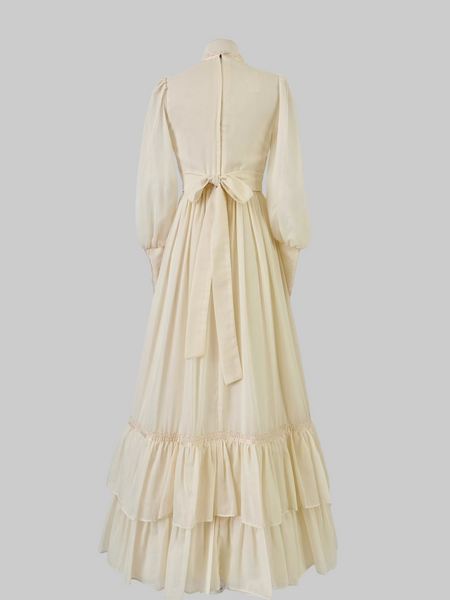 1970s Gunne Sax vintage cream gown with an embroidered yoke, lace-trimmed high neckline with bow tie, faux-pearl buttoned bodice, billowy sleeves with zippered cuffs, waist ties, and a ruffled hem.