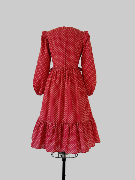 1970s Candi Jones prairie dress in crimson calico print featuring a wide lace collar, ivory rose buttons, self-tie waist bow, ruffled shoulders, and a tiered hem.