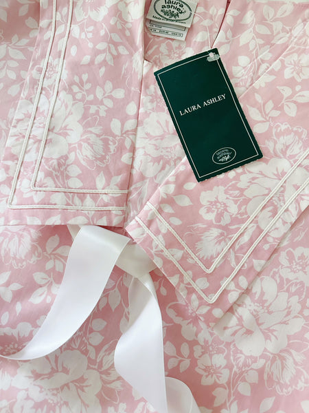 A close-up of a pink and white floral print dress by Laura Ashley, featuring white piping, a white satin tie belt, and the brand's dark green hangtag.