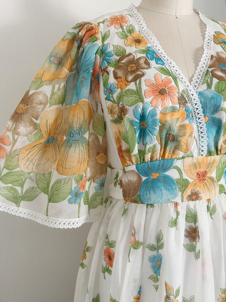 A close up of vintage 1970s white dress with a colorful floral print, featuring blue, orange, and yellow flowers with green leaves. The dress has a lace-trimmed V-neckline and wide, short flutter sleeves with white lace trim.