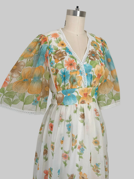 A vintage 1970s floral print dress . The dress features a white base with a colorful floral pattern in orange, blue, yellow, and green, and has white lace trim along the V-neckline and sleeve cuffs.