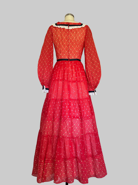 Back View of Mid-1970s crimson red calico prairie maxi dress with off-white lace trim, navy ribbon accents, long sleeves, and a tiered full-length skirt.