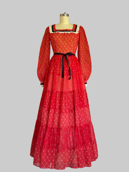 Mid-1970s crimson red calico prairie maxi dress with off-white lace trim, navy ribbon accents, long sleeves, and a tiered full-length skirt.