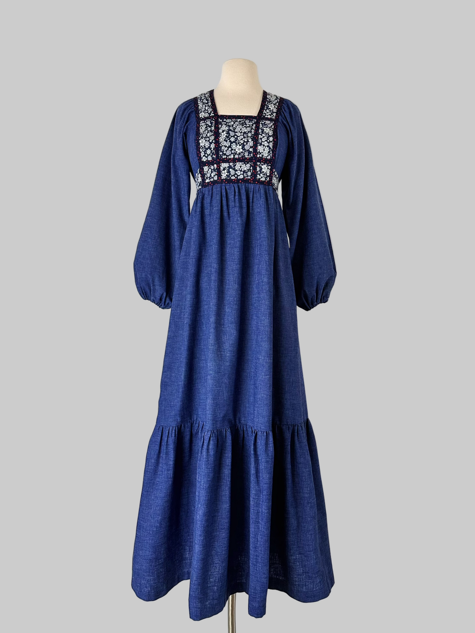 Vintage early-to-mid 1970s prairie dress in denim blue with a white floral calico yoke, long peasant sleeves, matching waist ties, and a tiered ruffle skirt. Features a soft woven cotton-poly fabric and back zipper closure.