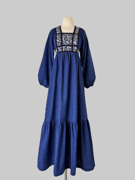 Vintage early-to-mid 1970s prairie dress in denim blue with a white floral calico yoke, long peasant sleeves, matching waist ties, and a tiered ruffle skirt. Features a soft woven cotton-poly fabric and back zipper closure.