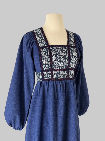 Vintage early-to-mid 1970s prairie dress in denim blue with a white floral calico yoke, long peasant sleeves, matching waist ties, and a tiered ruffle skirt. Features a soft woven cotton-poly fabric and back zipper closure.