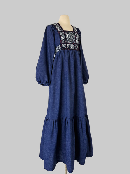 Vintage early-to-mid 1970s prairie dress in denim blue with a white floral calico yoke, long peasant sleeves, matching waist ties, and a tiered ruffle skirt. Features a soft woven cotton-poly fabric and back zipper closure.