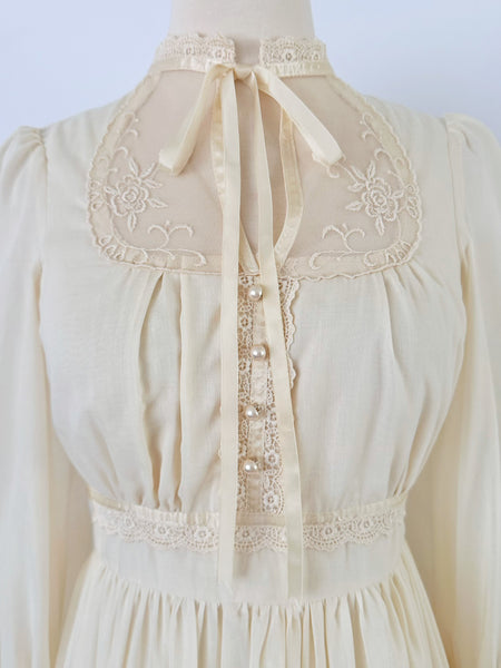 1970s Gunne Sax vintage cream gown with an embroidered yoke, lace-trimmed high neckline with bow tie, faux-pearl buttoned bodice, billowy sleeves with zippered cuffs, waist ties, and a ruffled hem.