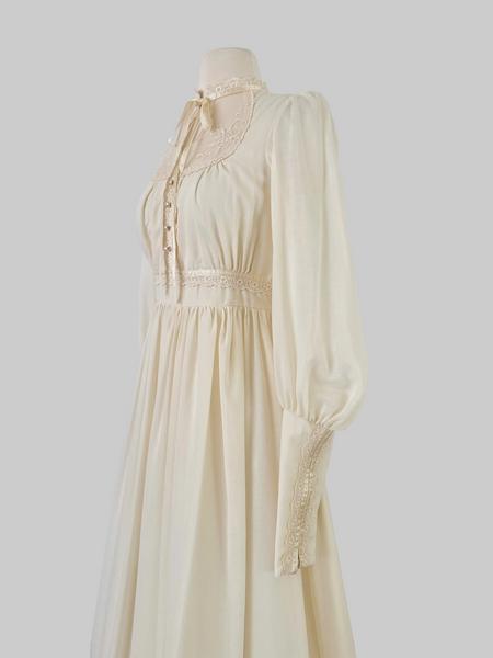 1970s Gunne Sax vintage cream gown with an embroidered yoke, lace-trimmed high neckline with bow tie, faux-pearl buttoned bodice, billowy sleeves with zippered cuffs, waist ties, and a ruffled hem.